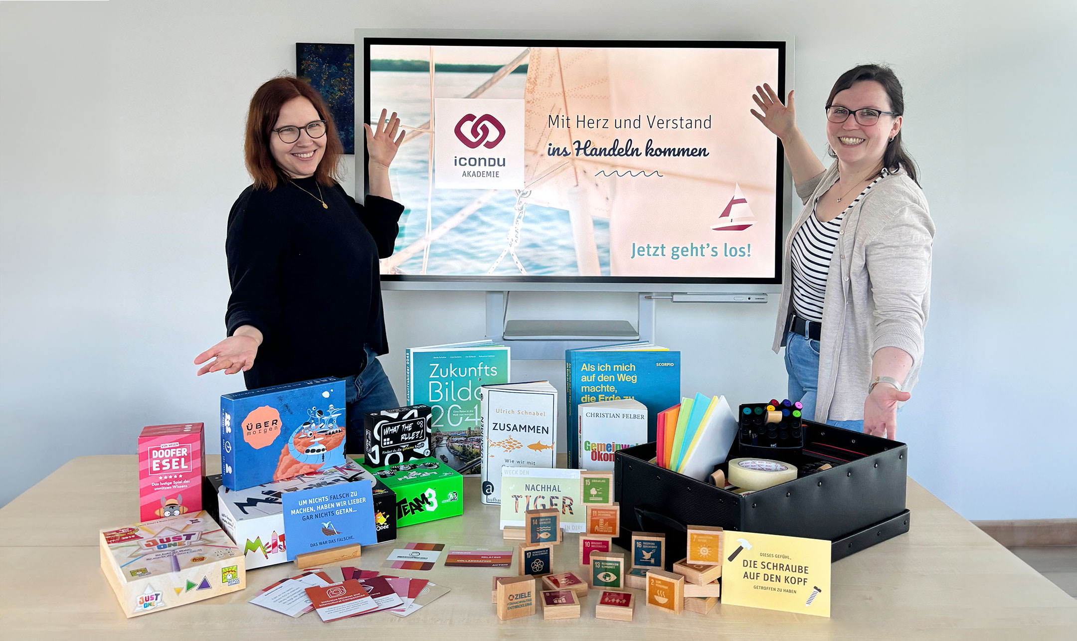 Barbara Holzner und Carina Heyer präsentieren freudig eine Sammlung von Brettspielen und Workshopmaterial.